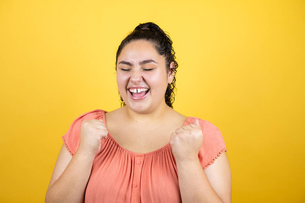 Young beautiful woman with curly hair over isolated yellow background very happy and excited making winner gesture with raised arms, smiling and screaming for success. - Photo, image