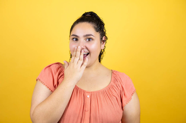 Young beautiful woman with curly hair over isolated yellow background with her hand over her mouth and surprised - Photo, Image
