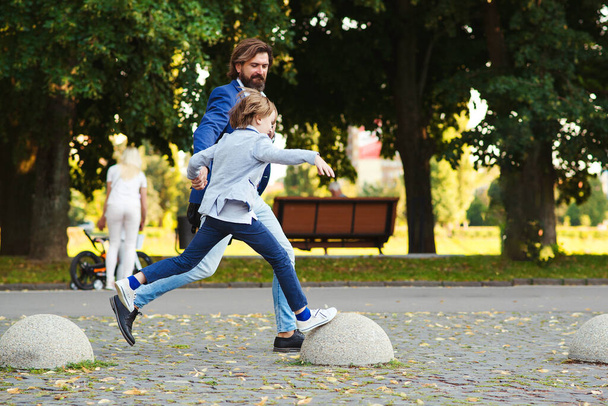 Ευτυχισμένος πατέρας και γιος διασκεδάζουν μαζί σε μια βόλτα. Μοντέρνος μπαμπάς και παιδί τρέχουν στο καλοκαιρινό πάρκο. Γενειοφόρος άντρας και αγόρι που φοράνε μοδάτα κοστούμια. Γονιός και παιδί πάνε σπίτι μετά το σχολείο.. - Φωτογραφία, εικόνα