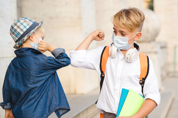 Children with face mask going back to school. New greeting style. Kids bumping elbows outdoors. Social distance. Education during coronavirus pandemic. - Photo, Image