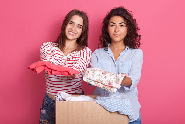 Two friends girls holding clothes in hands, showing shirts to camera, preparing box with clothing for charity, ladies posing against rose background. - Foto, afbeelding