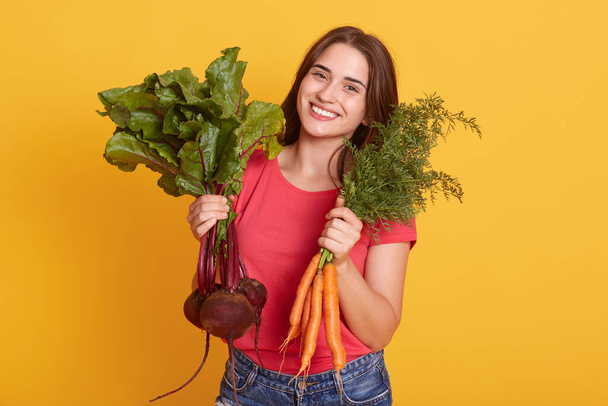 Portrait of smiling female gardener with carrot and beetroots in hands, looking smiling at camera, girl vegetarian wearing casual red t shirt against yellow wall. - Fotó, kép