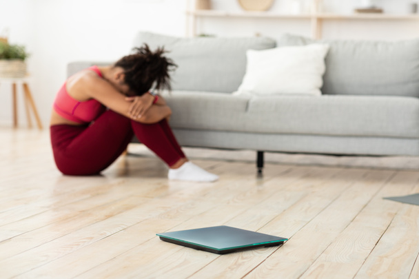 Черная женщина плачет сидя рядом с весом весы после взвешивания дома - Фото, изображение