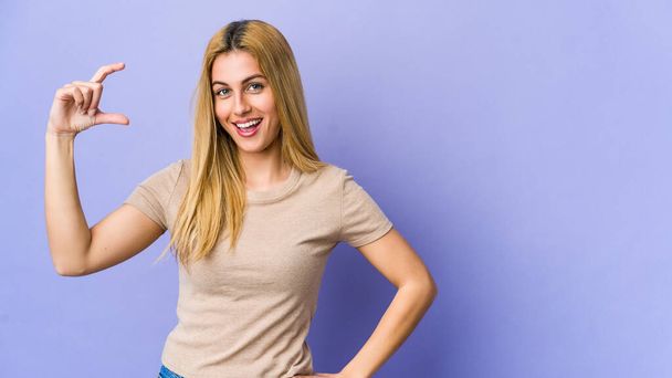 Mladá blondýna žena izolované na fialovém pozadí drží něco málo s prsty, usmívá a sebevědomě. - Fotografie, Obrázek