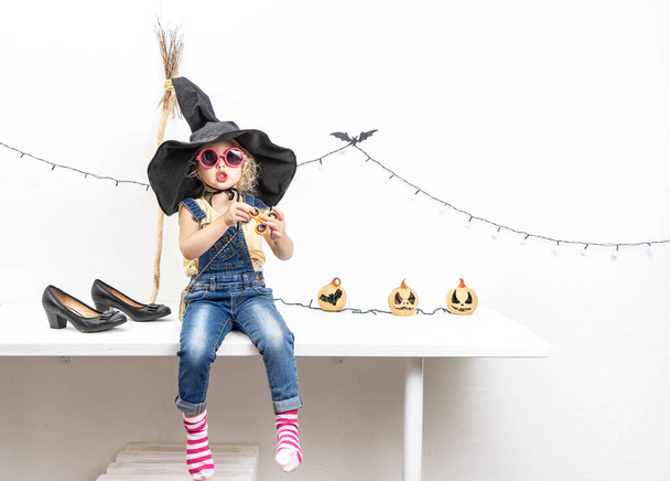 Happy Halloween: A little girl sits on a table in a witch hat with a broom. - Foto, Bild