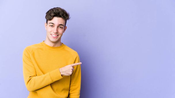 Young caucasian man isolated on purple background smiling and pointing aside, showing something at blank space. - Photo, Image