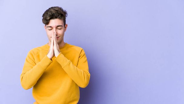 Jonge blanke man geïsoleerd op paarse achtergrond hand in hand bidden in de buurt van de mond, voelt vertrouwen. - Foto, afbeelding