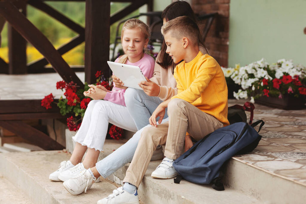 Μια ομάδα χαρούμενων εφήβων παιδιών μετά το σχολείο κάθονται στα σκαλιά και παίζουν σε μια ψηφιακή ταμπλέτα. Σύγχρονα παιδιά, ψηφιακές τεχνολογίες - Φωτογραφία, εικόνα