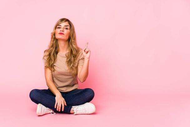 Young blonde caucasian woman sitting on a pink studio showing number one with finger. - Foto, Imagen