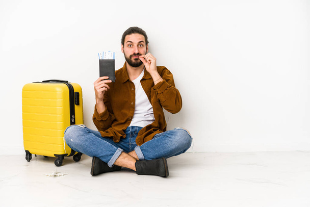 Giovane uomo caucasico seduto in possesso di un passaporto e una valigia isolata con le dita sulle labbra mantenendo un segreto. - Foto, immagini