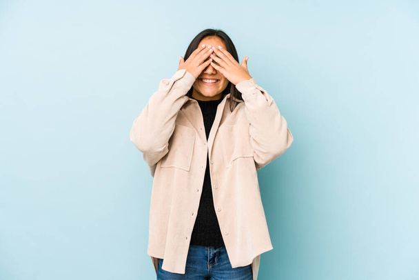 Jonge vrouw geïsoleerd op een blauwe achtergrond bedekt ogen met handen, glimlacht breed wachten op een verrassing. - Foto, afbeelding