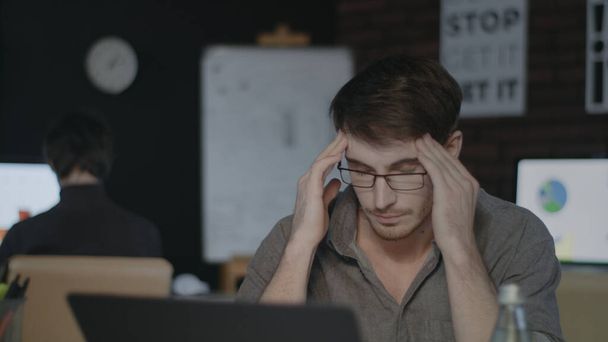 Κουρασμένος επιχειρηματίας που αγγίζει το κεφάλι στο σκοτεινό γραφείο. Θλιβερός άνθρωπος εργάζεται σκληρά για φορητό υπολογιστή. - Φωτογραφία, εικόνα