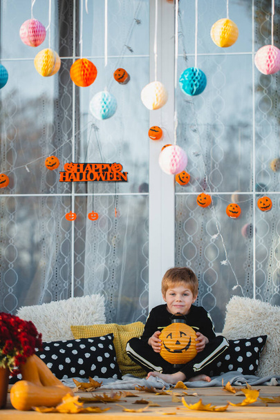 A beautiful child in a skeleton costume posing with a paper lantern in the form of a pumpkin. Decorations for the celebration of Halloween on the doorstep. Cozy veranda in a country house decorated for the holiday - Фото, зображення