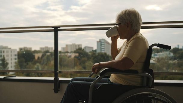 Oudere grijze vrouw met bril die koffie drinkt in de rolstoel op het balkon - Foto, afbeelding