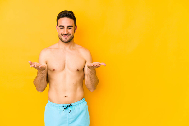 Young caucasian man with swimsuit isolated on yellow background Young caucasian man with trholding something with palms, offering to camera. - Fotó, kép