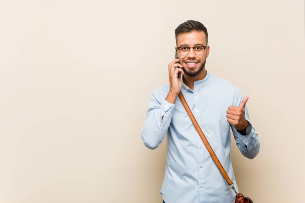 joven mixta raza asiático hombre de negocios sosteniendo un teléfono sonriendo y levantando el pulgar hasta - Foto, Imagen