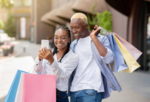 Boni, Geschenke und Bargeld nach dem Einkauf. Glückliches Paar mit vielen Farbbeuteln schaut aufs Smartphone - Foto, Bild