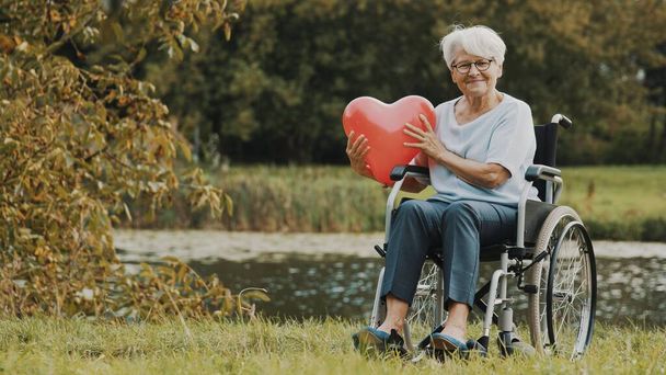 старшая женщина в инвалидной коляске возле реки держит шарик в форме сердца - Фото, изображение