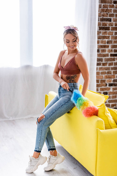 Young housewife in jeans cleaning sofa with dust brush  - Fotografie, Obrázek