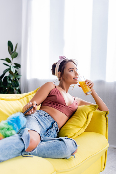 Selective focus of housewife drinking orange juice while holding dust brush on couch  - 写真・画像