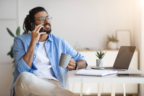 Indian Freelancer Guy Talking On Cellphone And Drinking Coffee At Home Office - Foto, imagen