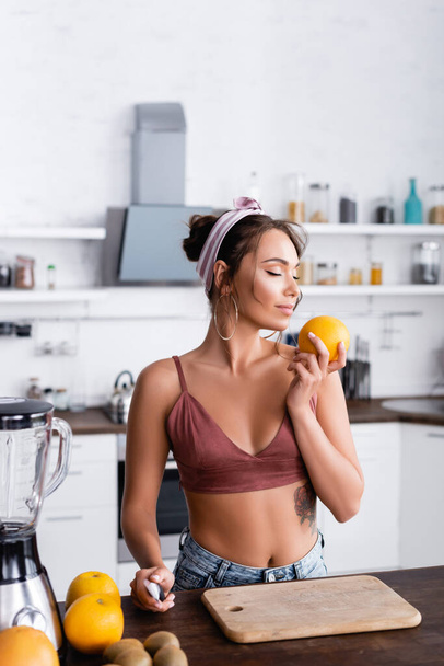 Selective focus of housewife smelling orange near fruits and blender in kitchen  - Fotografie, Obrázek