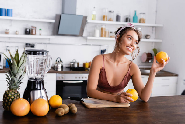Selective focus of young woman holding oranges near cutting board, fruits and blender at home  - Foto, Bild