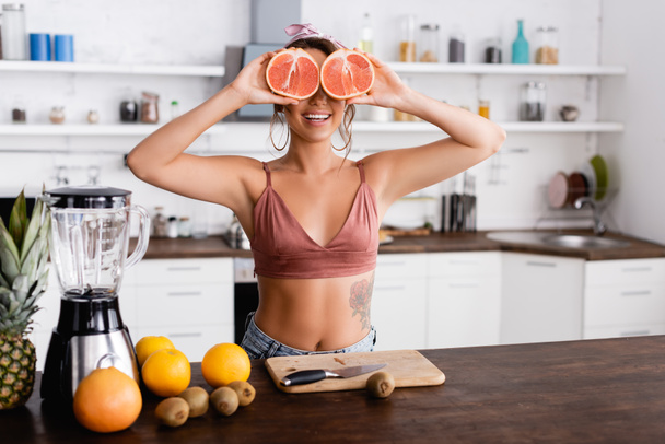 Selective focus of young woman holding halves of grapefruit near eyes while cooking smoothie in kitchen  - Photo, Image