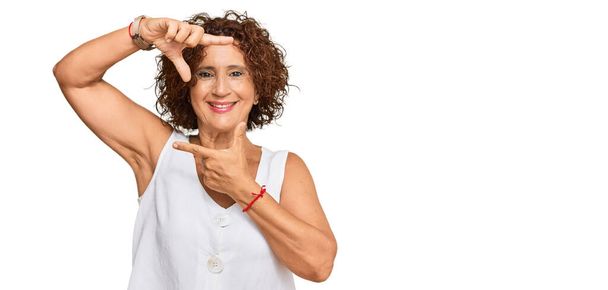 Mooie middelbare leeftijd volwassen vrouw dragen casual wit shirt glimlachen maken frame met handen en vingers met gelukkig gezicht. creativiteit en fotografie.  - Foto, afbeelding