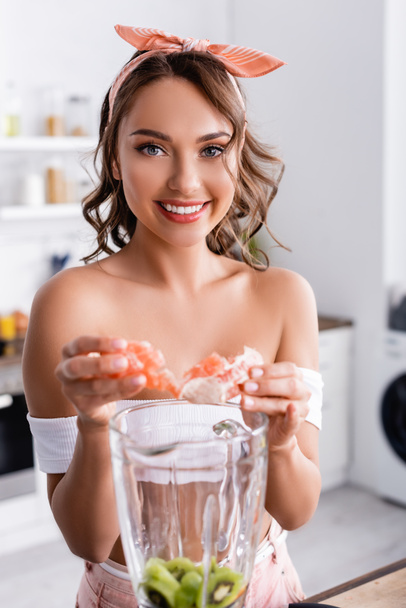 Selective focus of housewife looking at camera while adding fruits in blender  - Foto, immagini