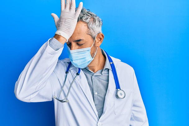 Middle age grey-haired man wearing doctor uniform and medical mask surprised with hand on head for mistake, remember error. forgot, bad memory concept.  - Фото, изображение