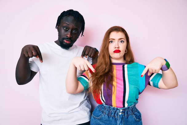 Interracial couple wearing casual clothes pointing down looking sad and upset, indicating direction with fingers, unhappy and depressed.  - Valokuva, kuva