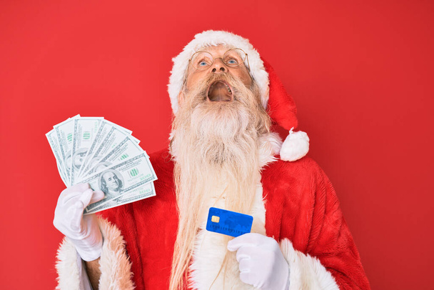 Staruszek w kostiumie Świętego Mikołaja trzymający dolary i kartę kredytową wściekły i wściekły krzyczący sfrustrowany i wściekły, krzyczący ze złości patrzący w górę.  - Zdjęcie, obraz