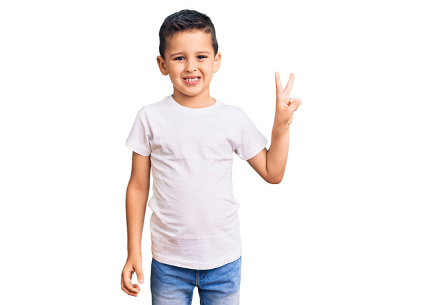 Petit garçon mignon portant un t-shirt blanc décontracté souriant avec un visage heureux clin d'oeil à la caméra faisant signe de victoire. numéro deux.  - Photo, image