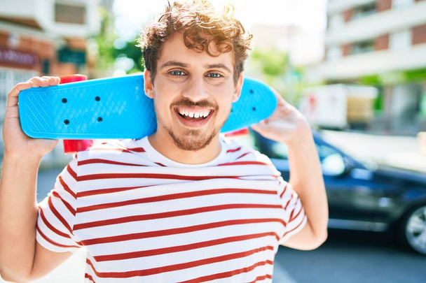 Junger kaukasischer Skater Mann lächelt glücklich und hält Skate zu Fuß auf der Straße der Stadt. - Foto, Bild