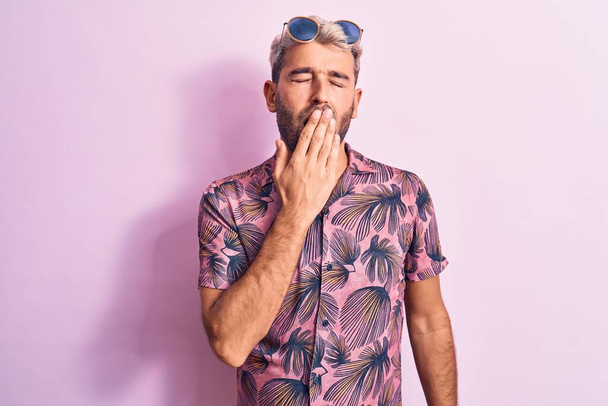 Przystojny blondyn na wakacjach ubrany w luźną koszulę i okulary przeciwsłoneczne na różowym tle znudzony ziewaniem zmęczony przykrywaniem ust dłonią. Niepokój i senność. - Zdjęcie, obraz