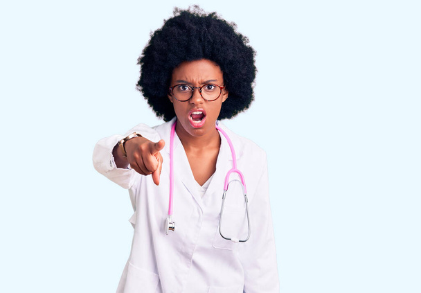 Fiatal afro-amerikai nő orvosi köpenyben és sztetoszkóppal, elégedetlen és frusztrált a kamerába, dühös és dühös rád.  - Fotó, kép