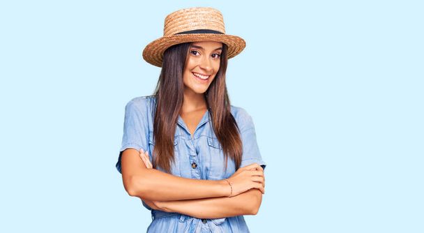 Νεαρή Ισπανίδα που φοράει καλοκαιρινό καπέλο χαμογελάει χαμογελώντας με σταυρωμένα τα χέρια κοιτάζοντας την κάμερα. θετικό πρόσωπο.  - Φωτογραφία, εικόνα