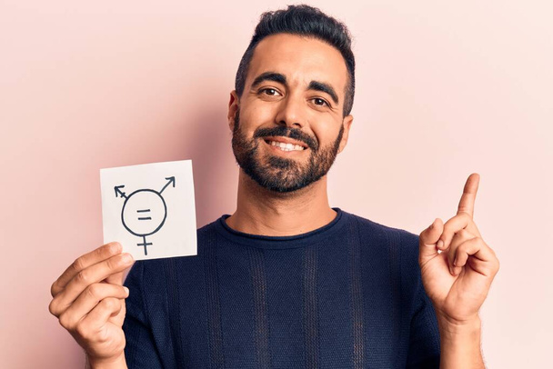 Молодой латиноамериканец, держащий в руках символ трансгендера, улыбаясь счастливо указывая рукой и пальцем в сторону  - Фото, изображение