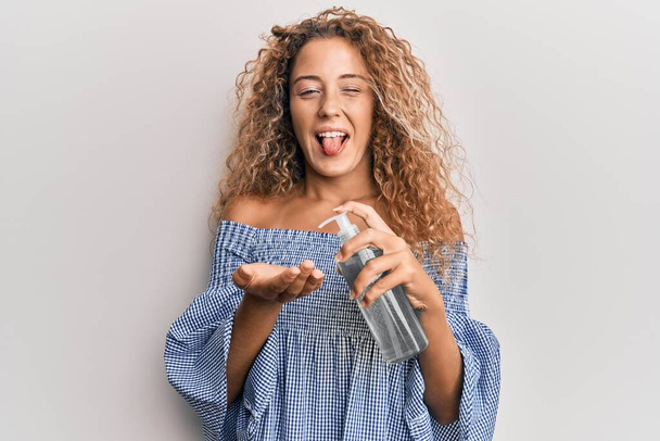 Belle adolescente caucasienne utilisant un gel désinfectant pour les mains langue collante heureux avec une expression drôle.  - Photo, image