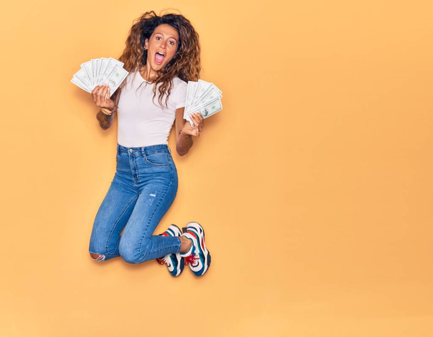 Young beautiful curly woman with tattoo surprised holding dollars banknotes. Jumping with smile on face and open mouth over isolated yellow background - Photo, Image