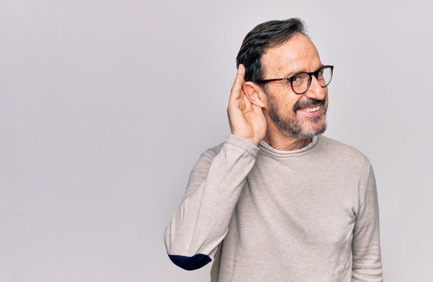 Middle age handsome man wearing casual sweater and glasses over isolated white background smiling with hand over ear listening and hearing to rumor or gossip. Deafness concept. - Photo, Image