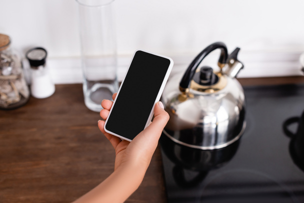 Περικοπή άποψη της γυναίκας κρατώντας smartphone με λευκή οθόνη κοντά στο βραστήρα στο φούρνο  - Φωτογραφία, εικόνα