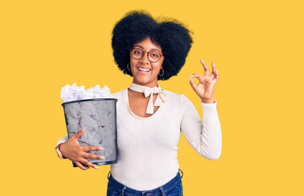 Joven afroamericana chica sosteniendo papelera llena de papeles arrugados haciendo ok signo con los dedos, sonriendo gesto amistoso excelente símbolo  - Foto, imagen