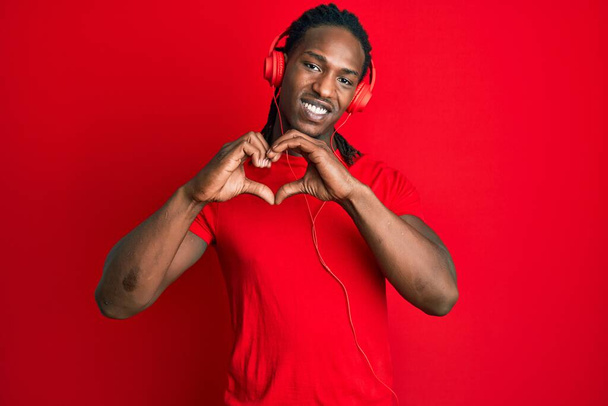 Homme afro-américain avec des tresses écoutant de la musique à l'aide d'écouteurs souriant en amour faisant forme de symbole de coeur avec les mains. concept romantique.  - Photo, image
