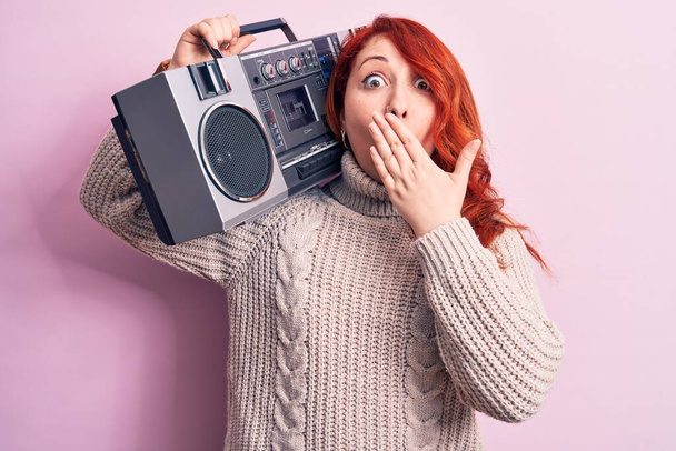 Mujer pelirroja hermosa escuchando música sosteniendo boombox vintage sobre fondo rosa cubriendo la boca con la mano, conmocionada y asustada por el error. Expresión sorprendida - Foto, Imagen