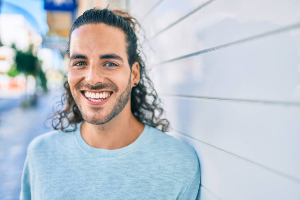 jonge latino man glimlachend gelukkig kijkend naar de camera leunend op de muur - Foto, afbeelding