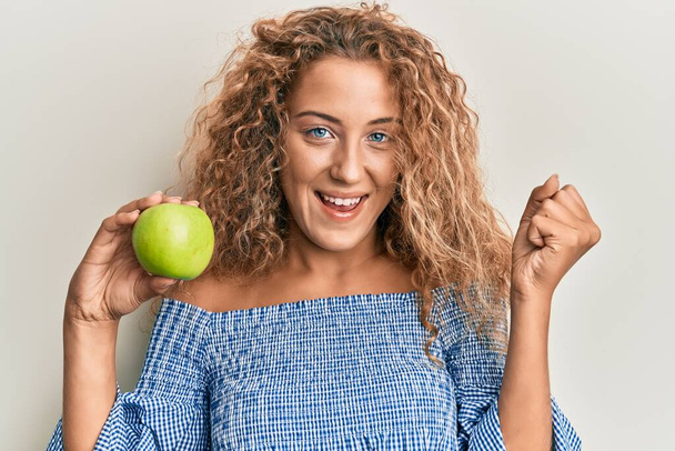 Beautiful caucasian teenager girl holding green apple screaming proud, celebrating victory and success very excited with raised arms  - Fotoğraf, Görsel