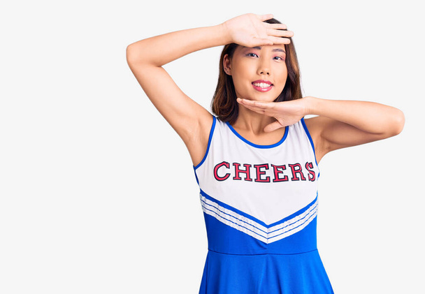 Giovane bella ragazza cinese indossa uniforme cheerleader sorridente allegra sbirciare una bua con le mani che mostrano il viso. sorpreso ed uscito  - Foto, immagini