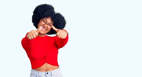 Νεαρή Αφρικάνα Αμερικανίδα που φοράει καθημερινά ρούχα και γυαλιά εγκρίνοντας να κάνει θετική χειρονομία με το χέρι, με τους αντίχειρες ψηλά χαμογελώντας και χαρούμενη για την επιτυχία. νικήτρια χειρονομία.  - Φωτογραφία, εικόνα